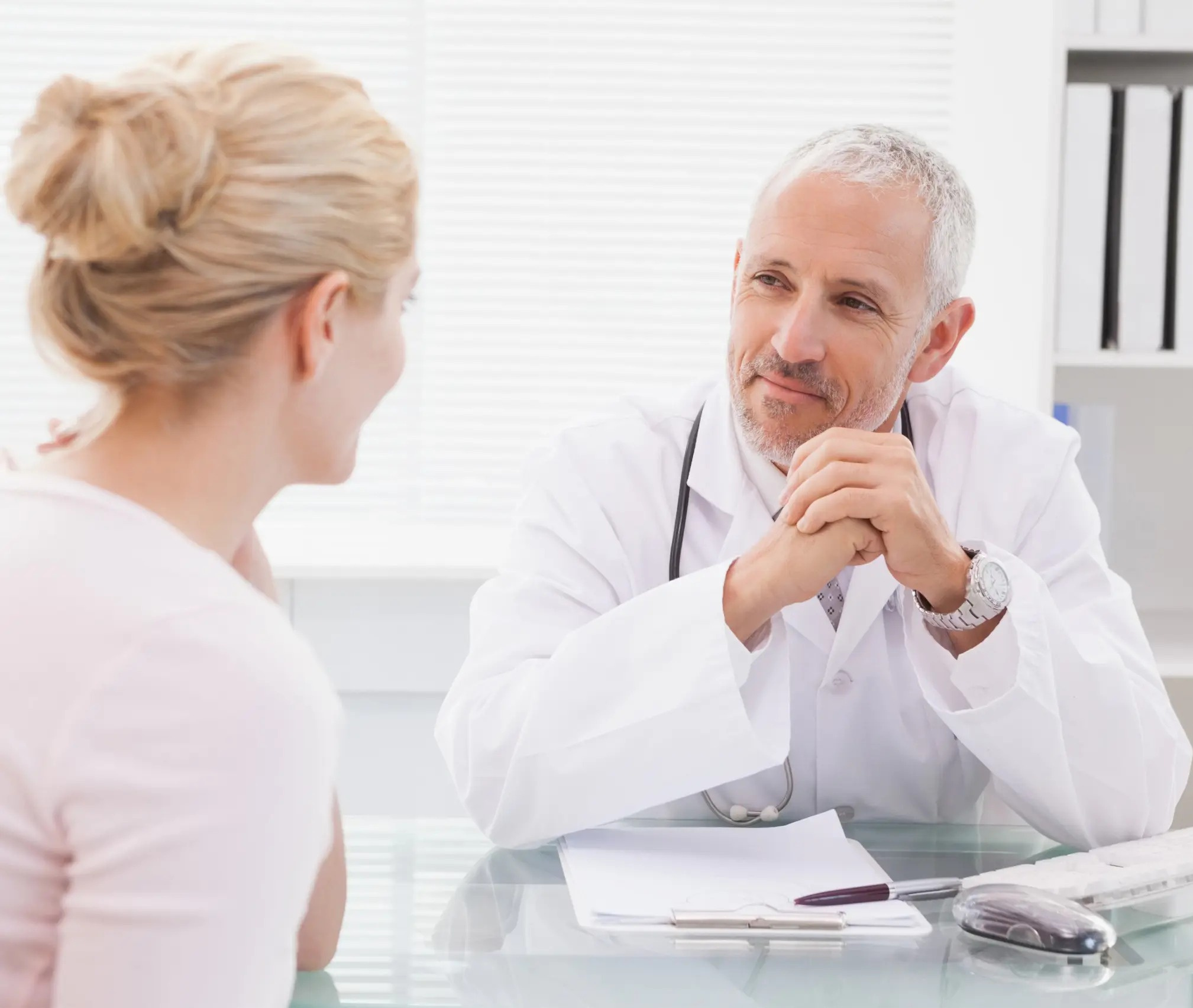 Eine Patientin und ein Arzt tauschen sich in einem modernen Behandlungsraum aus, während auf dem Tisch Unterlagen liegen.